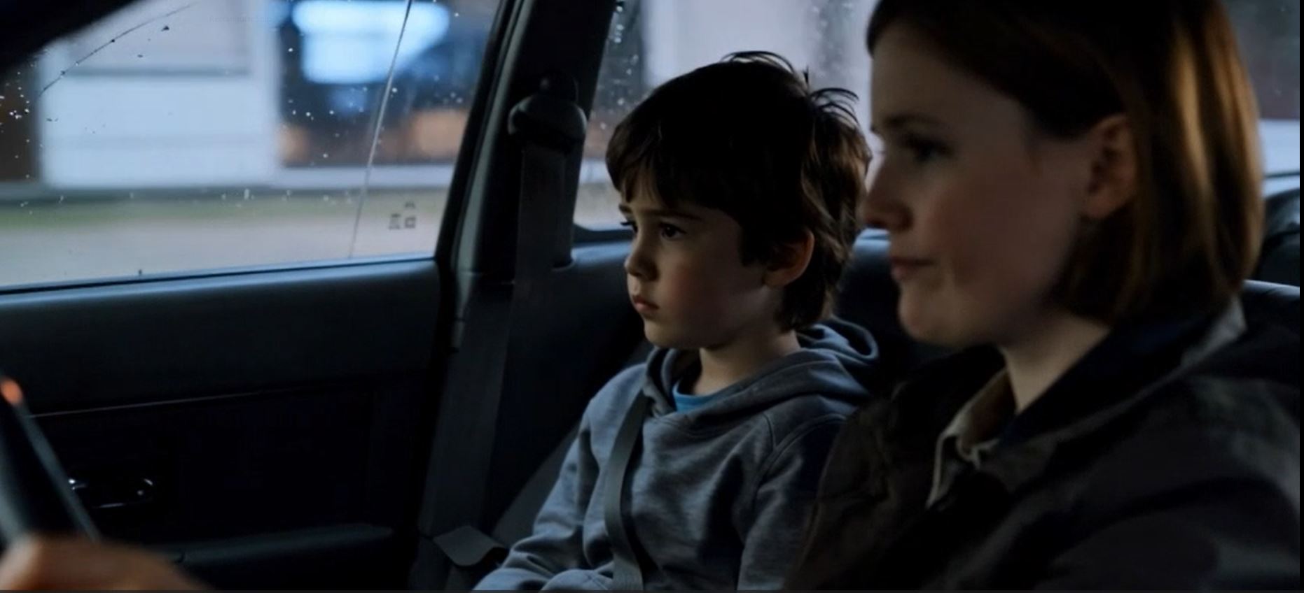View from the back of a family car after a junior football game, with a parent driving and a child in football kit beside them in soft afternoon light.