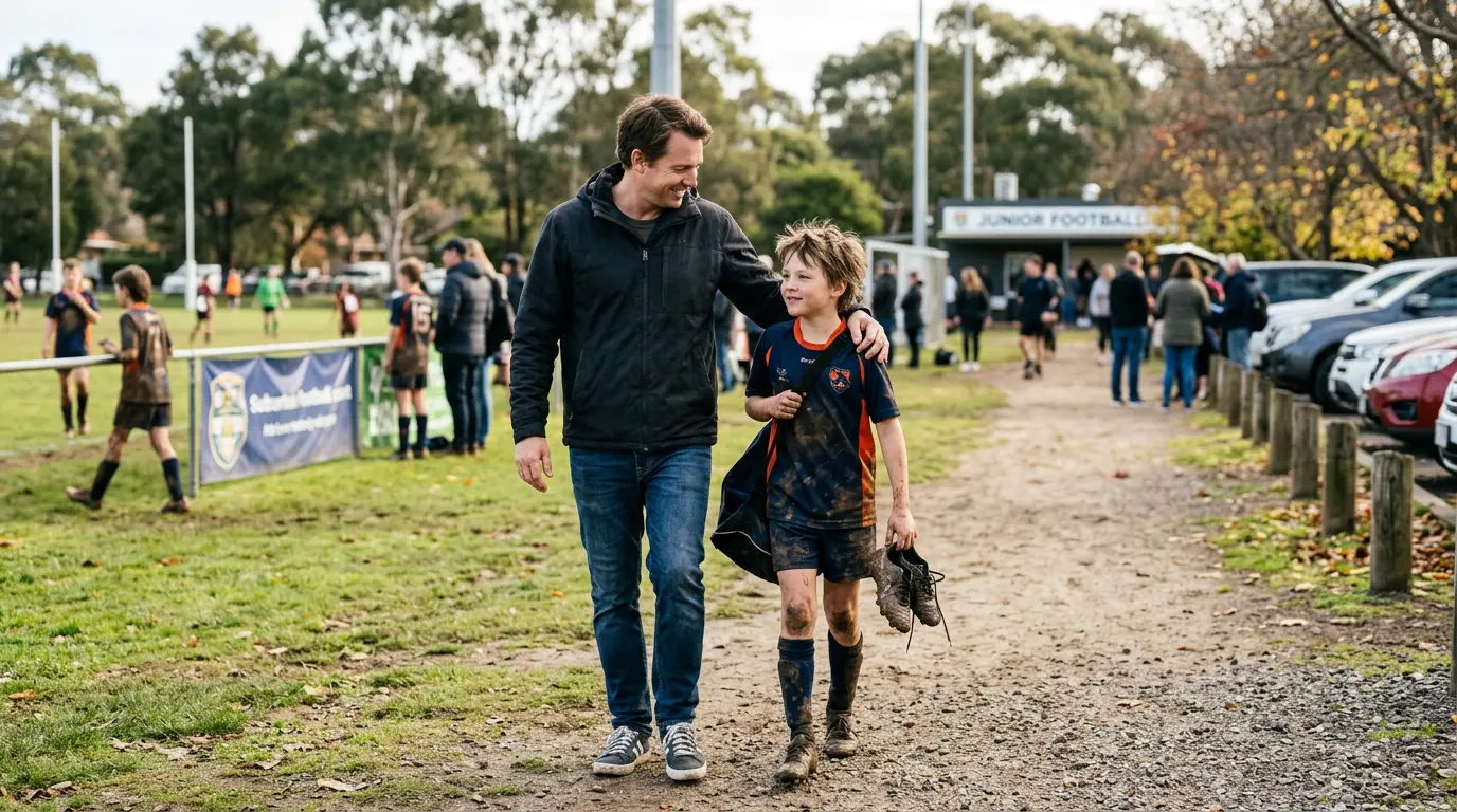 An Australian parent and their young child walk away from a community football pitch after a Saturday morning game. The parent's hand is on the kid's shoulder. Both are smiling, the parent looking sideways at the kid.