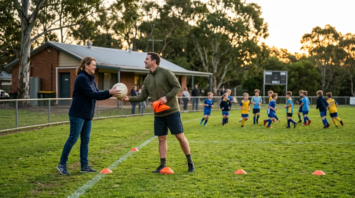 An Australian parent volunteer hands the coach a spare ball at training while the coach is setting up. A small everyday moment of support. Soft evening light at a community football ground.