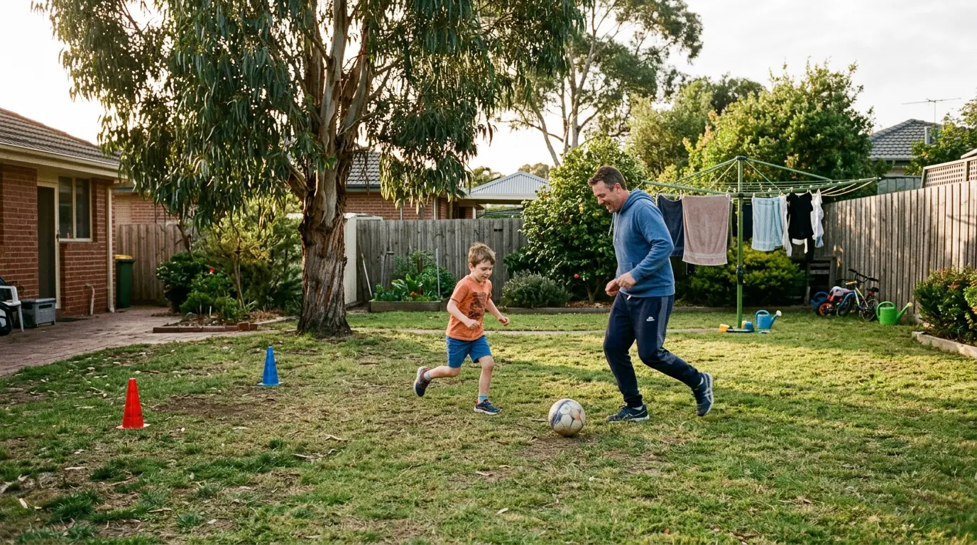 Two kids playing football in a suburban Australian backyard at golden hour, an old wooden chair used as a goal at one end and a lemon tree at the other, no adult or coach visible.